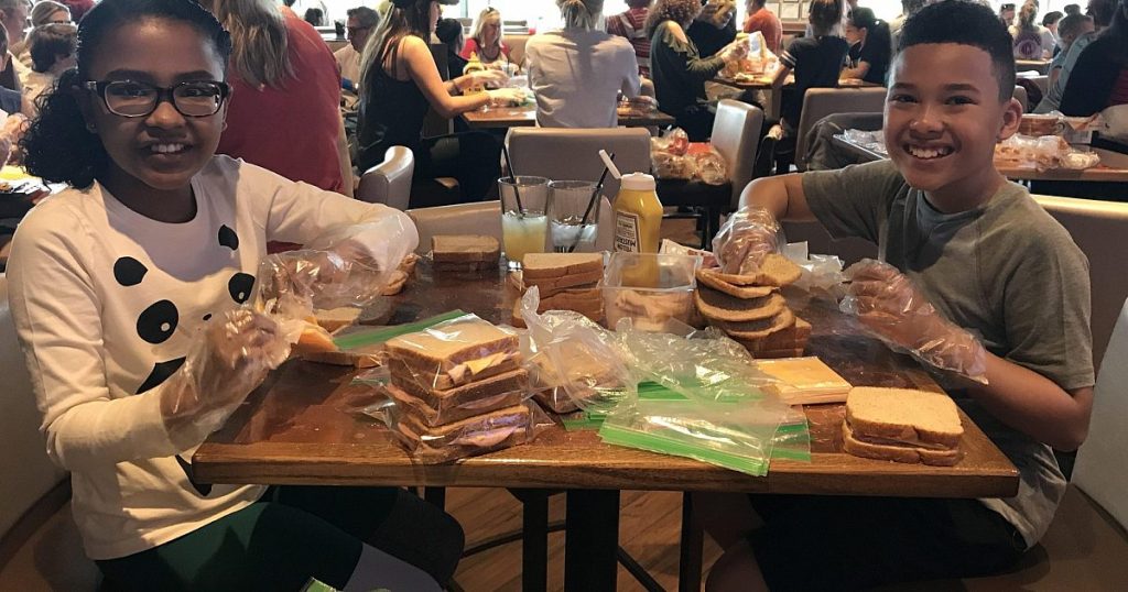 Two smiling children assembling bagged sandwiches at a table during a food charity event