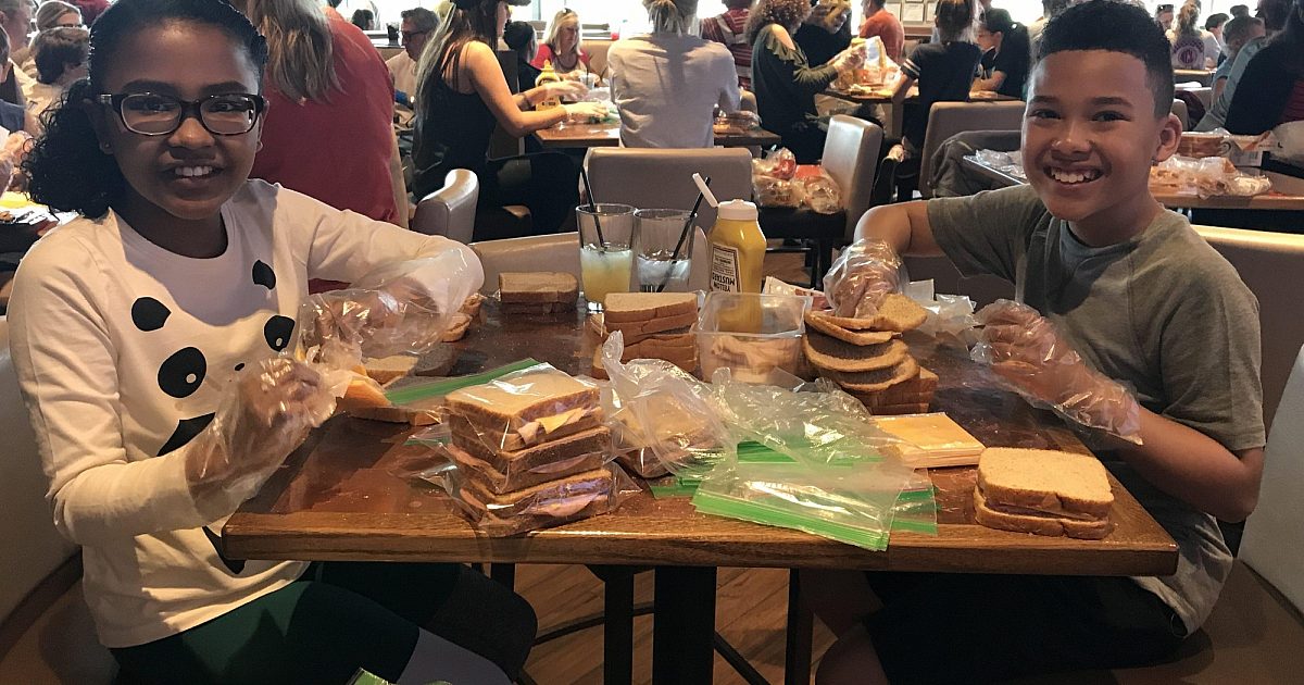 Two smiling children assembling bagged sandwiches at a table during a food charity event