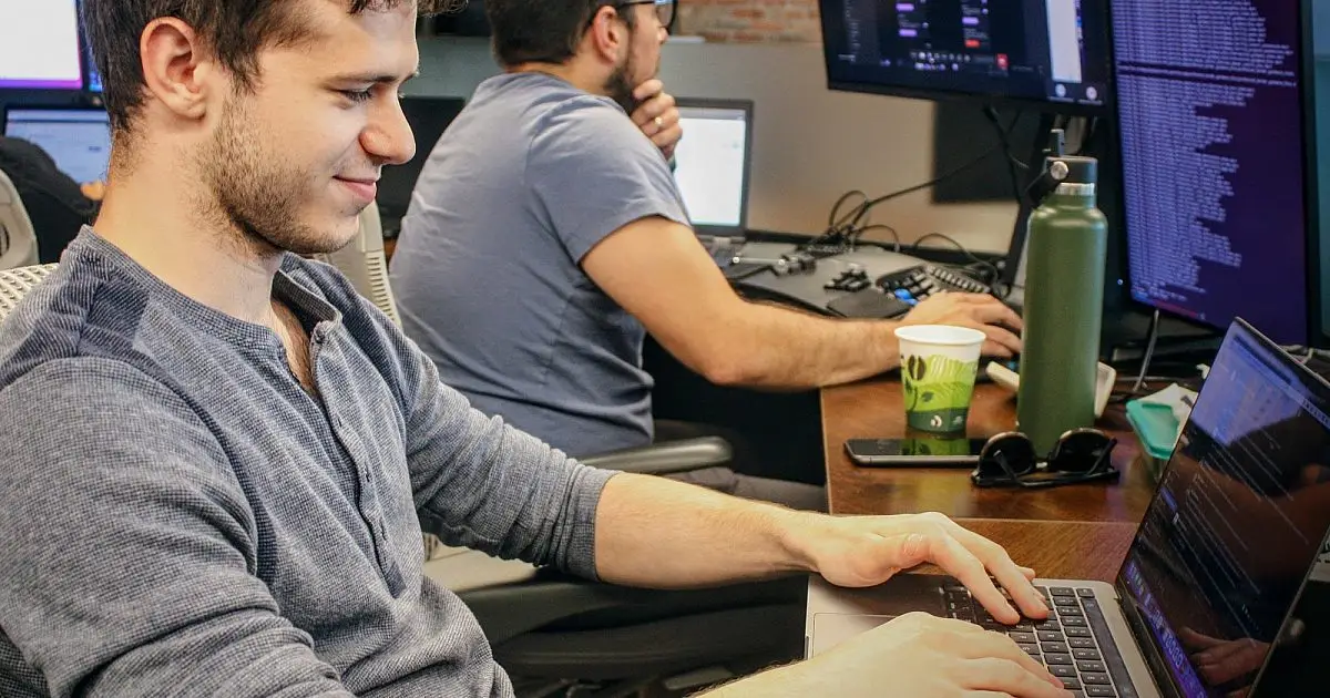 Two software engineers coding on laptops at a shared desk with monitors of code