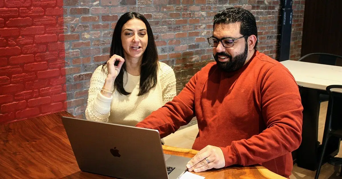 Two colleagues smiling and reviewing a laptop together at a table by a brick wall