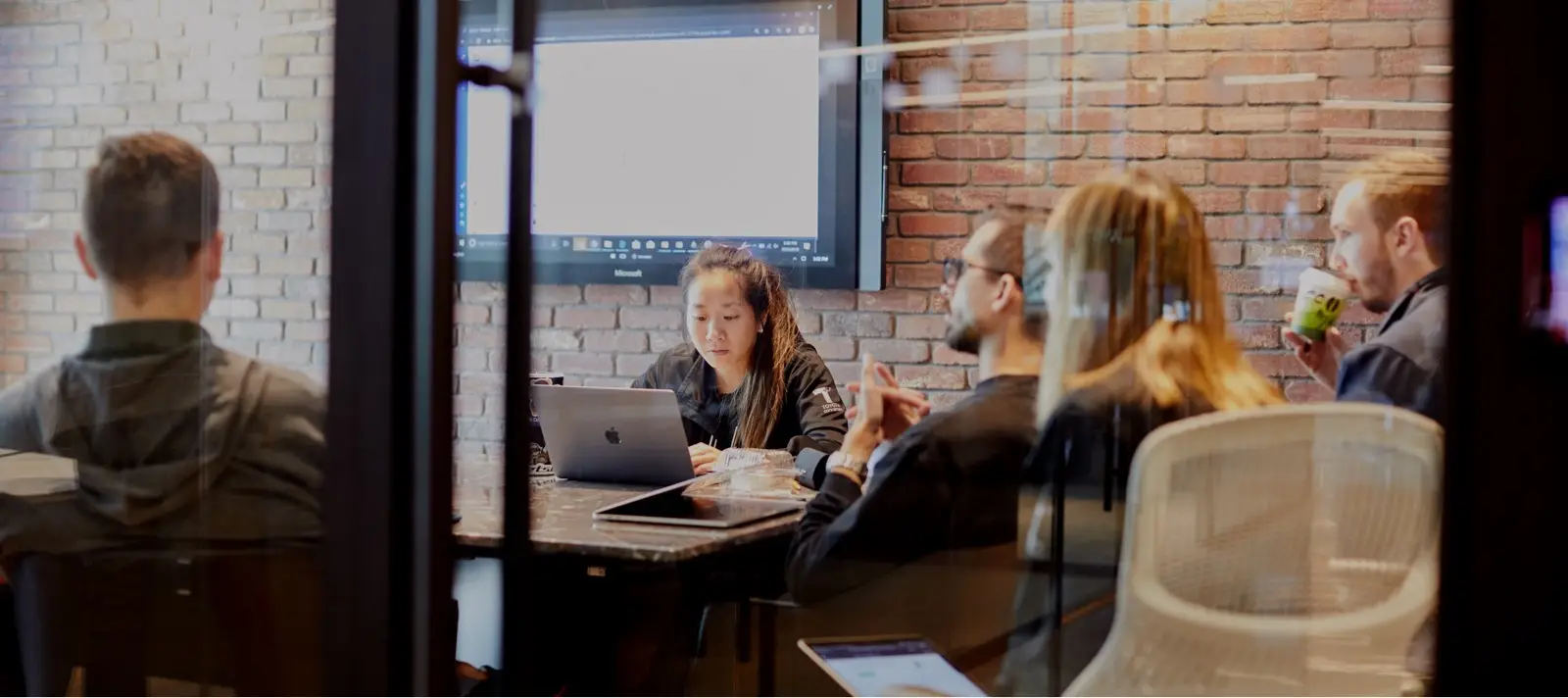 Team members collaborating around a laptop in a glass conference room with a wall display