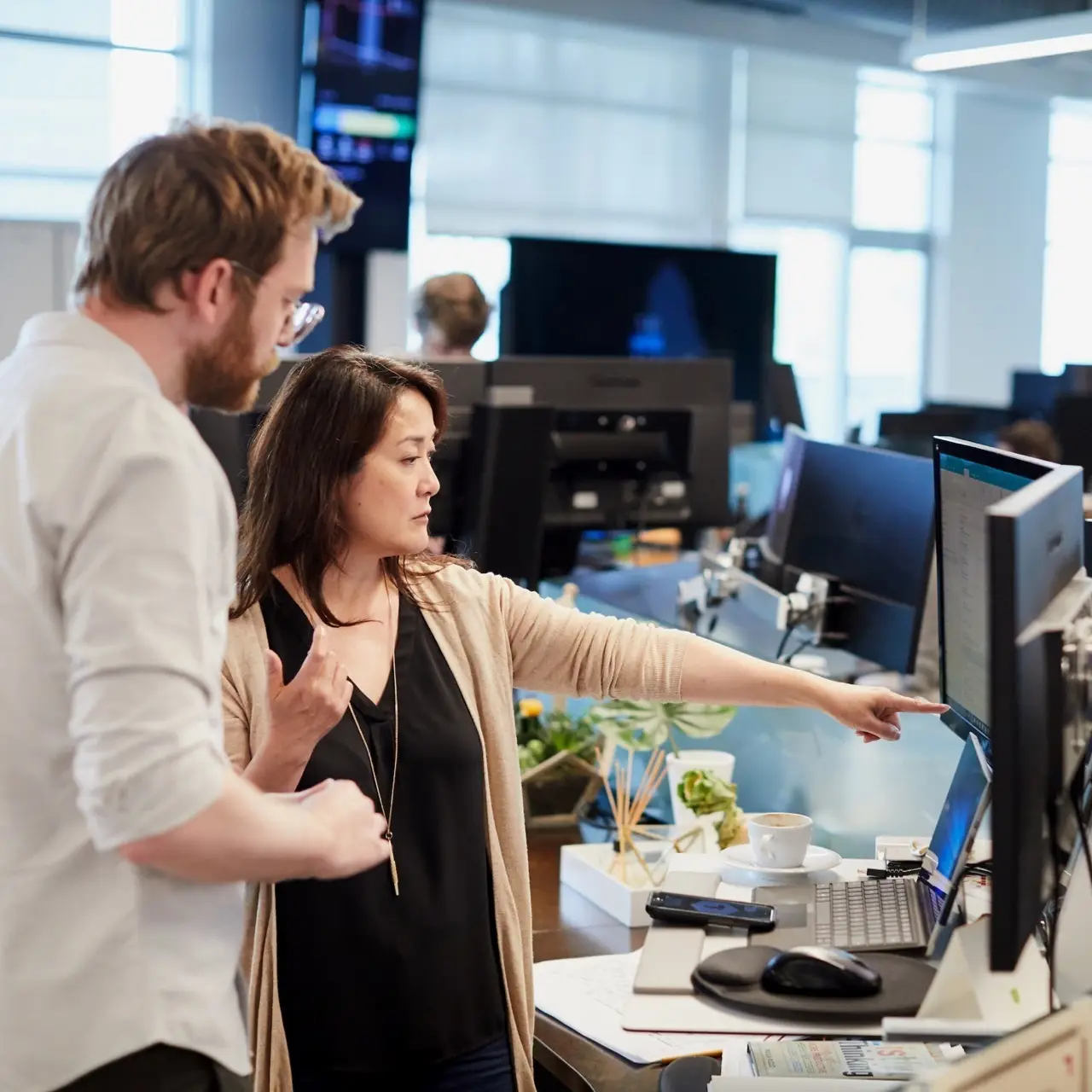 Two colleagues reviewing work together at a desk with multiple monitors in an open office