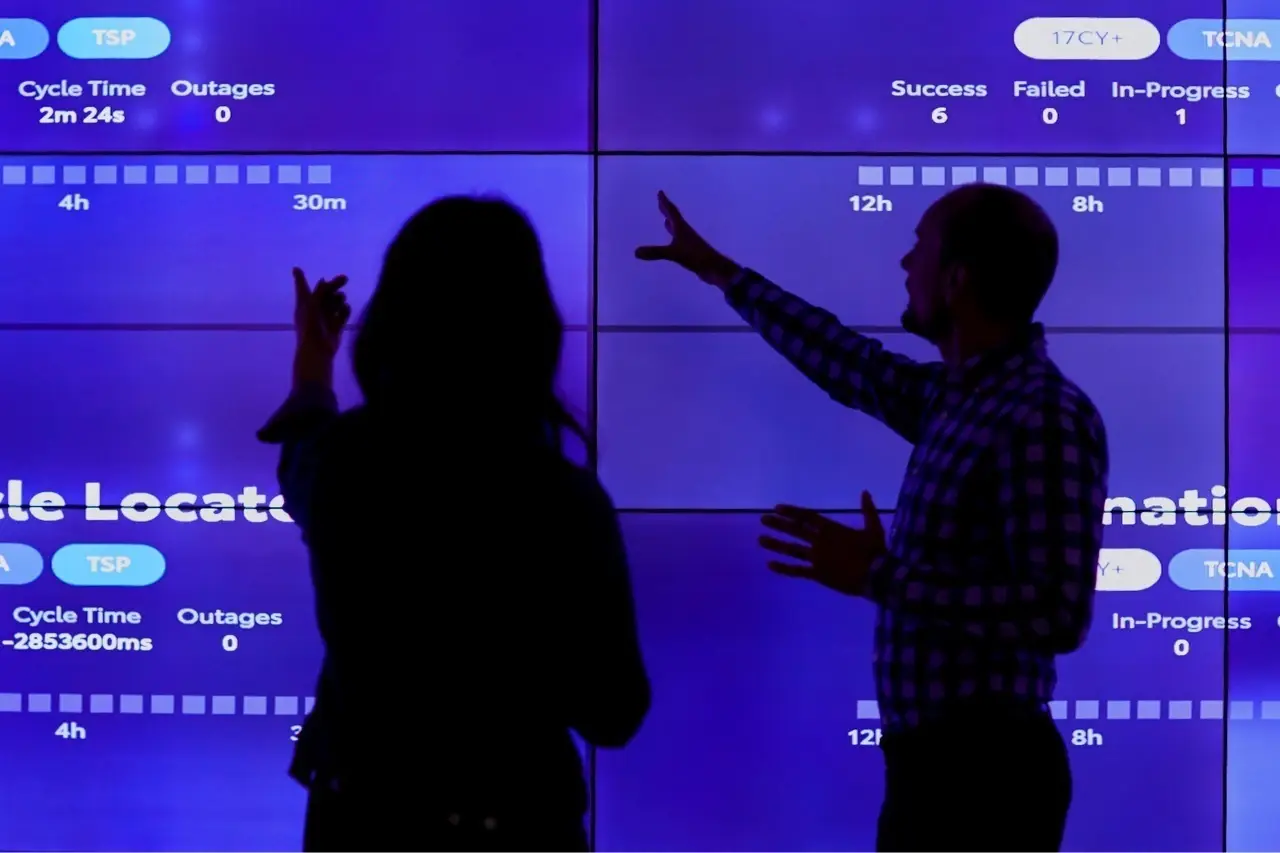 Two people silhouetted against a large data dashboard display showing performance metrics